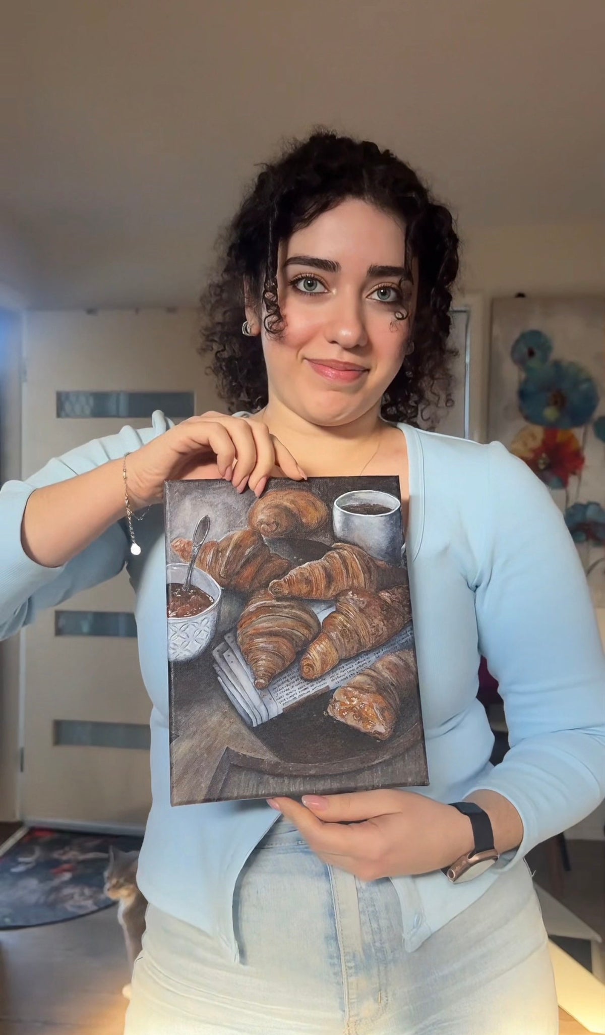 A woman with curly dark hair holds up a square canvas painting depicting croissants and a cup of coffee. She is wearing a light blue cardigan and white pants, standing in a home interior setting.